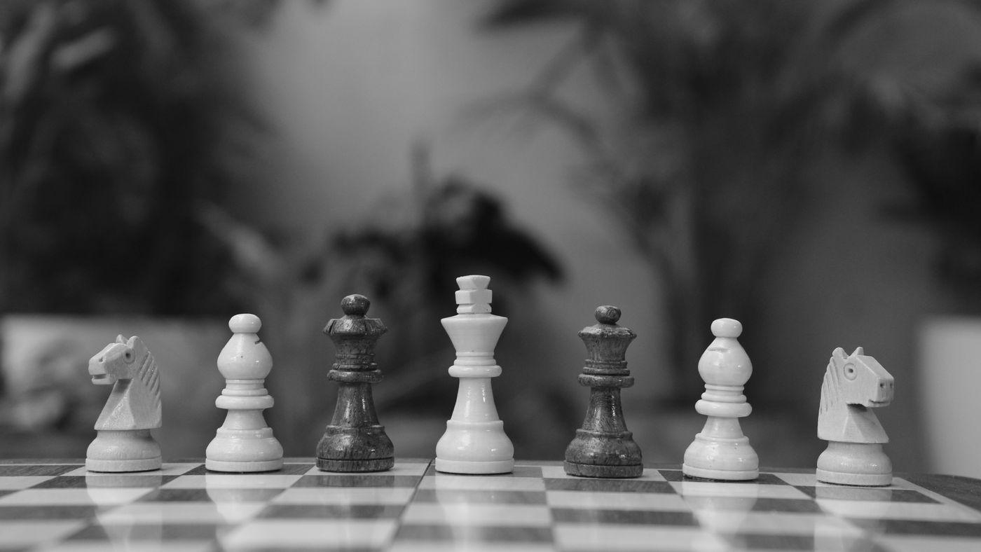 Monochrome image of chess pieces lined up on a chessboard with a blurred background.